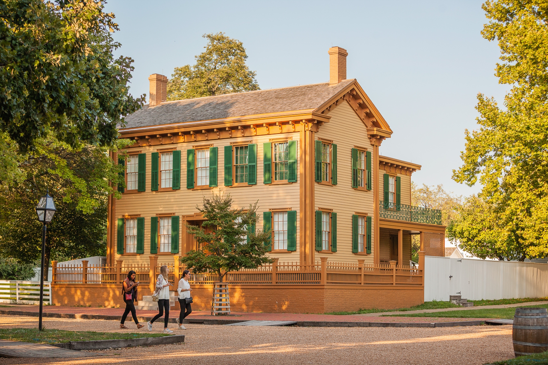 Picturesque historic two-story mustard-colored house in Illinois with green shutters, surrounded by lush trees, with pedestrians strolling by on a sunny day.