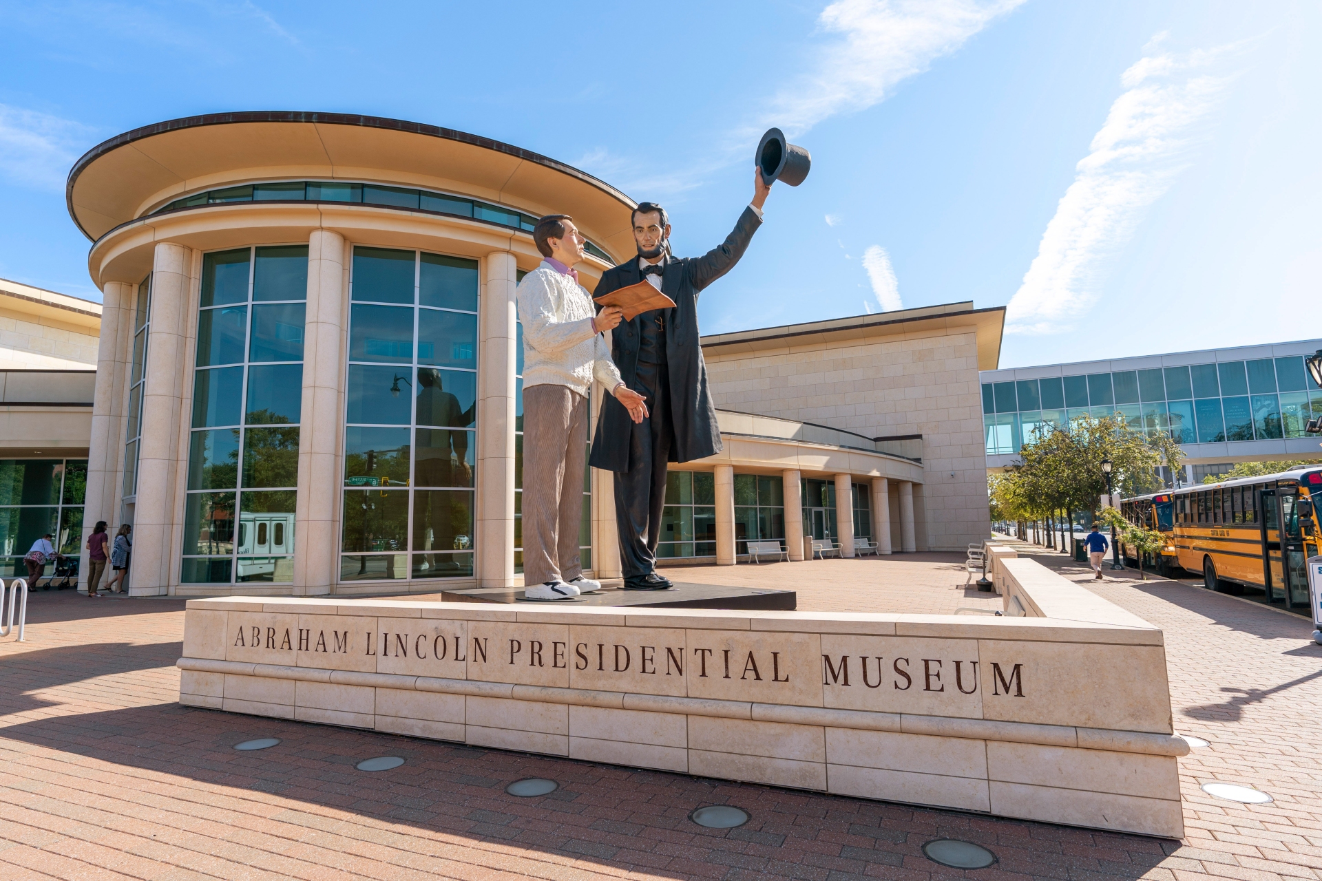 Sculpture of Abraham Lincoln greeting a modern visitor at the entrance of the Abraham Lincoln Presidential Museum in Illinois under a clear blue sky.