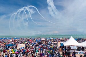 Crowds gather on a sunny day at a beachfront airshow, watching as aircraft perform precise aerial acrobatics against a backdrop of blue skies and scattered clouds.