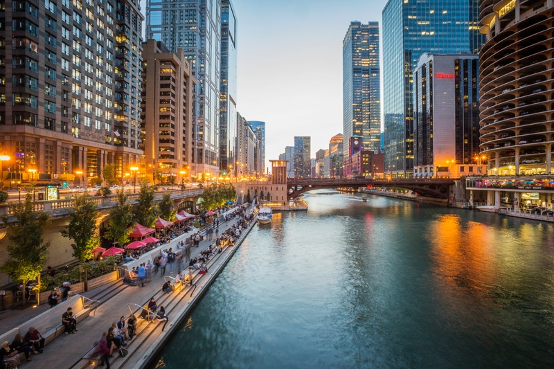 Bustling Illinois city riverwalk at dusk, with pedestrians enjoying the evening amidst high-rise buildings and a serene urban waterway.