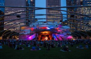 An evening outdoor concert at a modern bandshell with a large crowd enjoying the live performance under a vibrant, illuminated canopy against a backdrop of city skyscrapers.