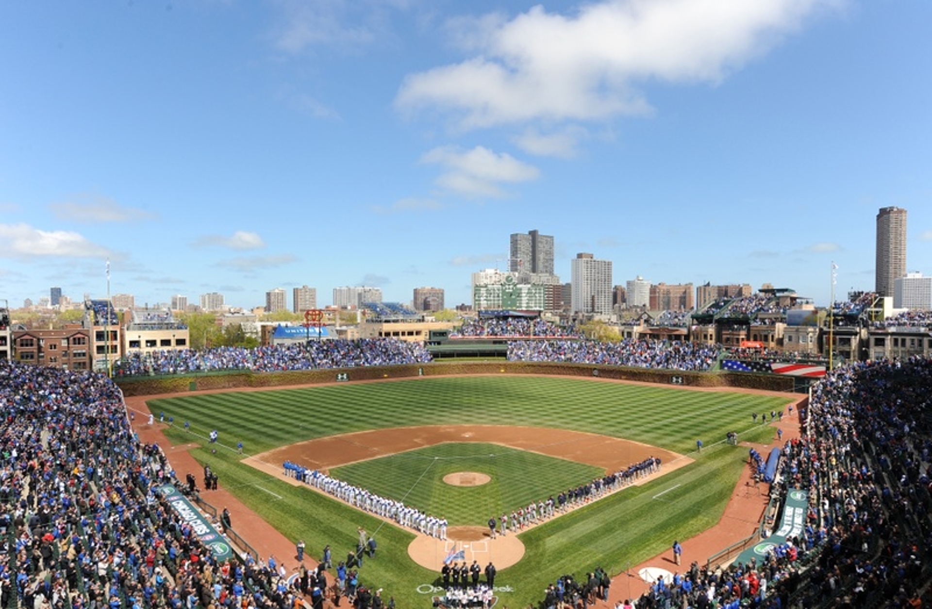 A panoramic view of a bustling baseball stadium filled with fans on a bright, sunny day in Illinois, with the city skyline looming in the background.