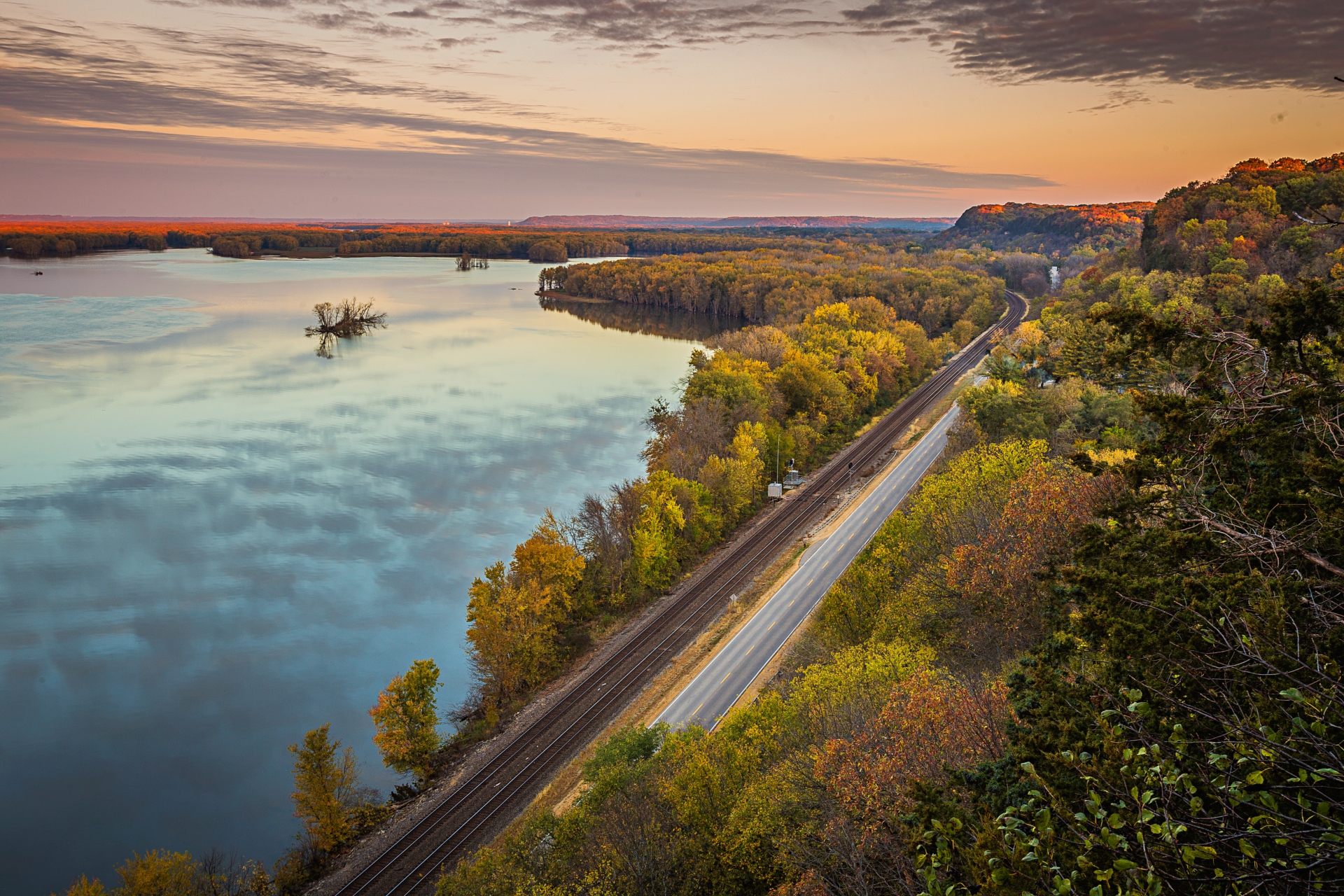 A serene autumn landscape in Illinois at sunset, featuring a tranquil lake beside a winding road and railway, with the foliage displaying vibrant fall colors.