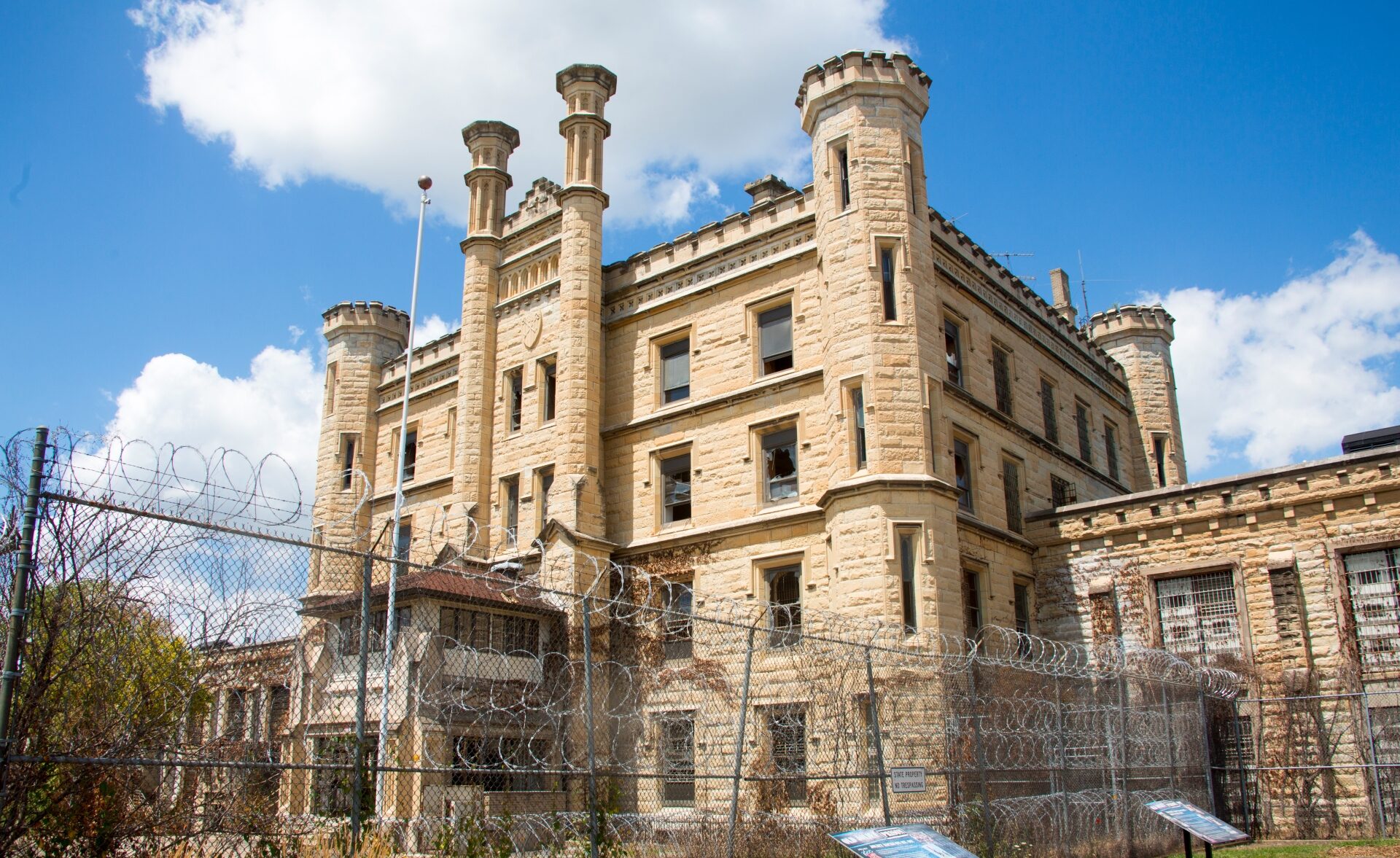 An imposing, historic stone building in Illinois, featuring castle-like architectural elements, with multiple chimneys and barred windows, secured behind a metal fence topped with rolls of barbed wire under a partly cloudy sky