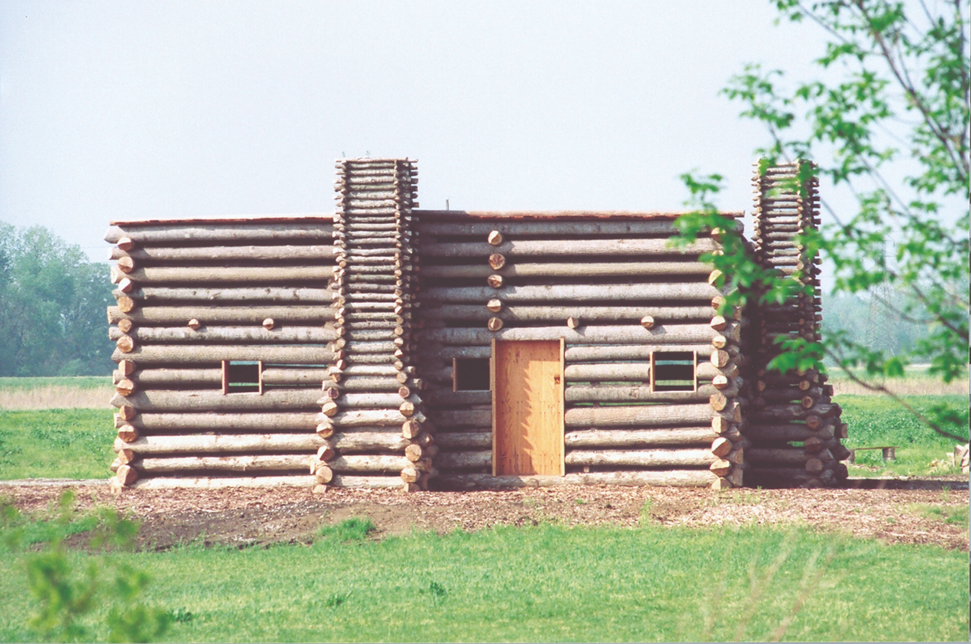 A sturdy log cabin with a single door and multiple small windows, set against a backdrop of open fields and a distant tree line under a hazy sky in Illinois.