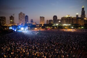 A vast crowd gathers at an outdoor music festival at dusk, with a brightly lit stage set against a city skyline.