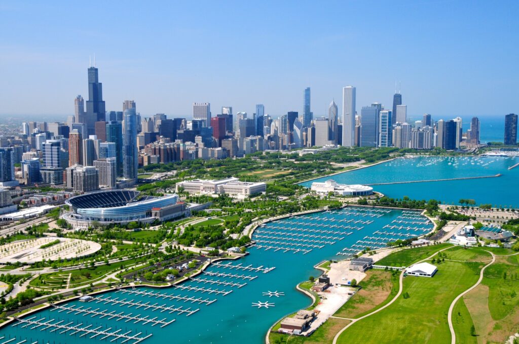 Aerial view of a sunlit Illinois chicago skyline with skyscrapers towering over parks and a marina along the shoreline of Lake Michigan.