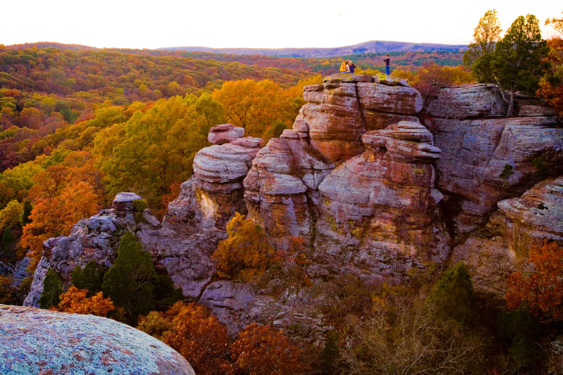 Autumn splendor from a high vantage point in Illinois: two individuals savor the view atop rugged sandstone cliffs, as a tapestry of fall foliage stretches into the horizon.