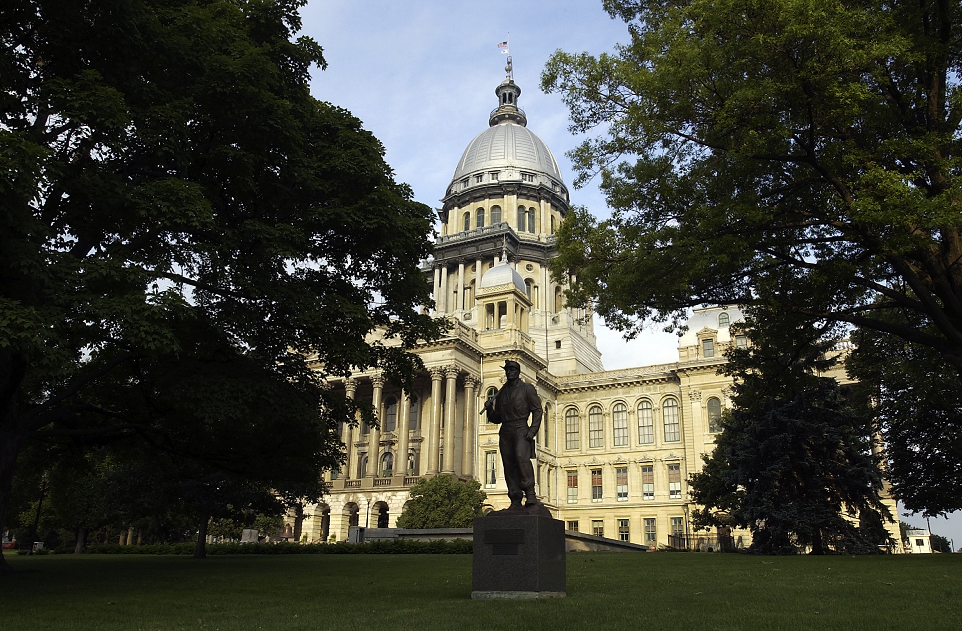 Statue standing in the foreground of the grand Illinois state capitol building adorned with a dome, surrounded by lush green trees.