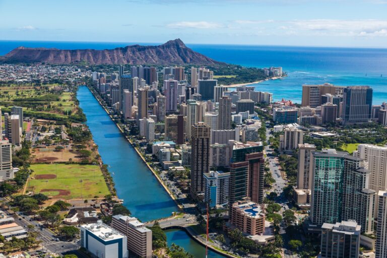 Aerial view of a vibrant cityscape with skyscrapers adjacent to a tranquil blue ocean, with a majestic mountain in the background under a clear sky.