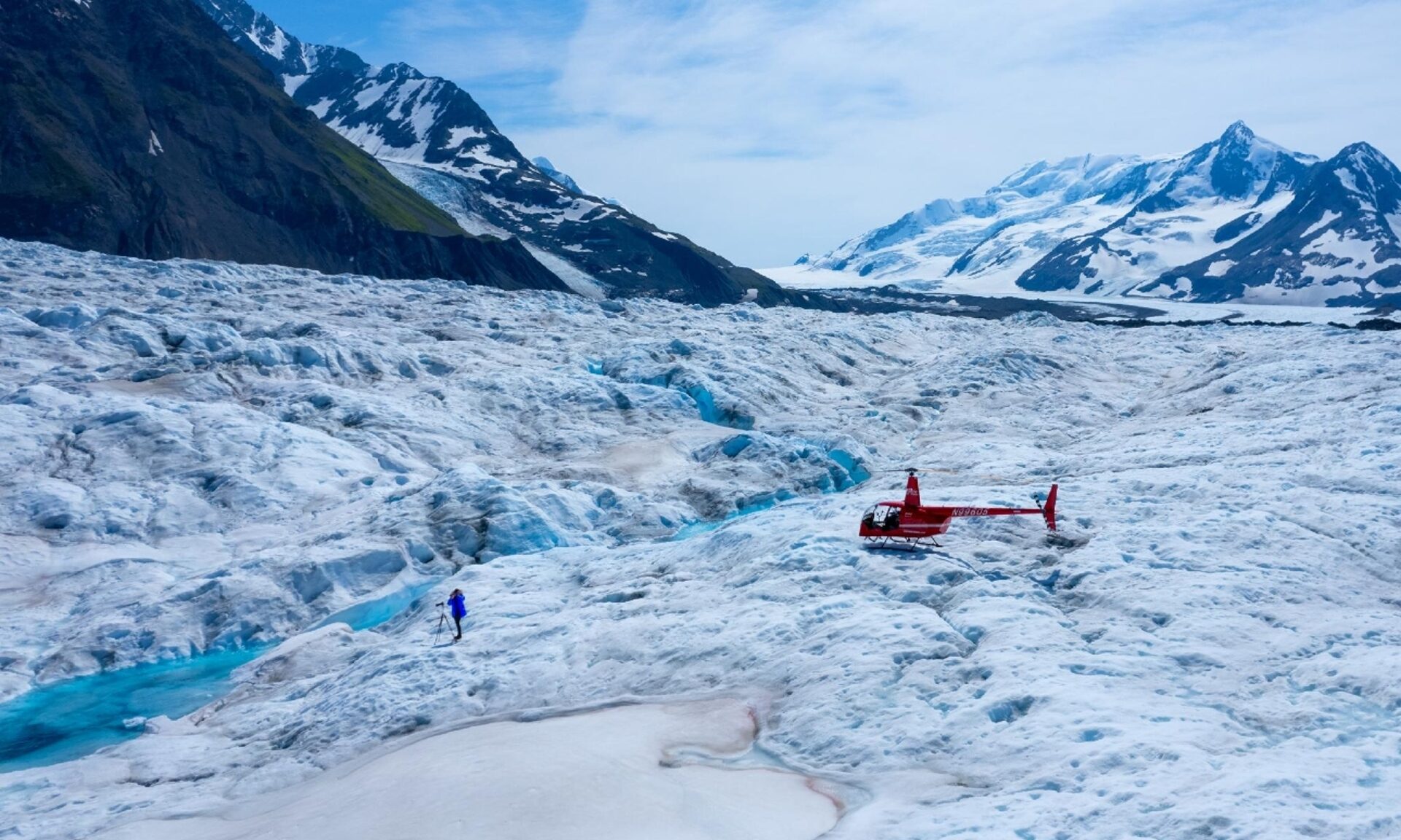 A red helicopter landing on a rugged glacier with a person nearby, highlighting the contrast between human technology and nature's raw beauty amidst snow-capped mountains.
