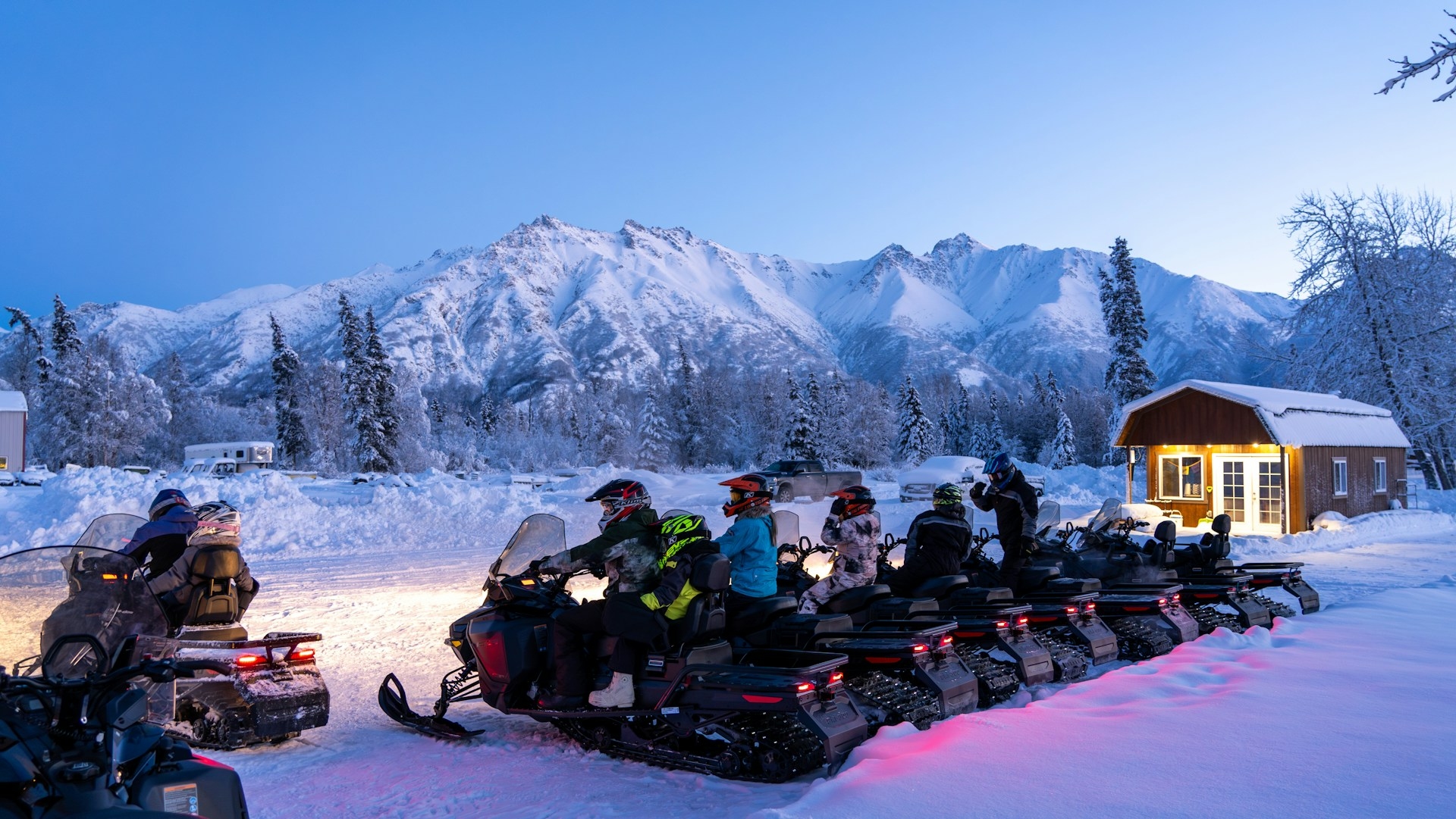 Group of snowmobile riders preparing for a twilight adventure in a snow-covered landscape with majestic mountains in the background.