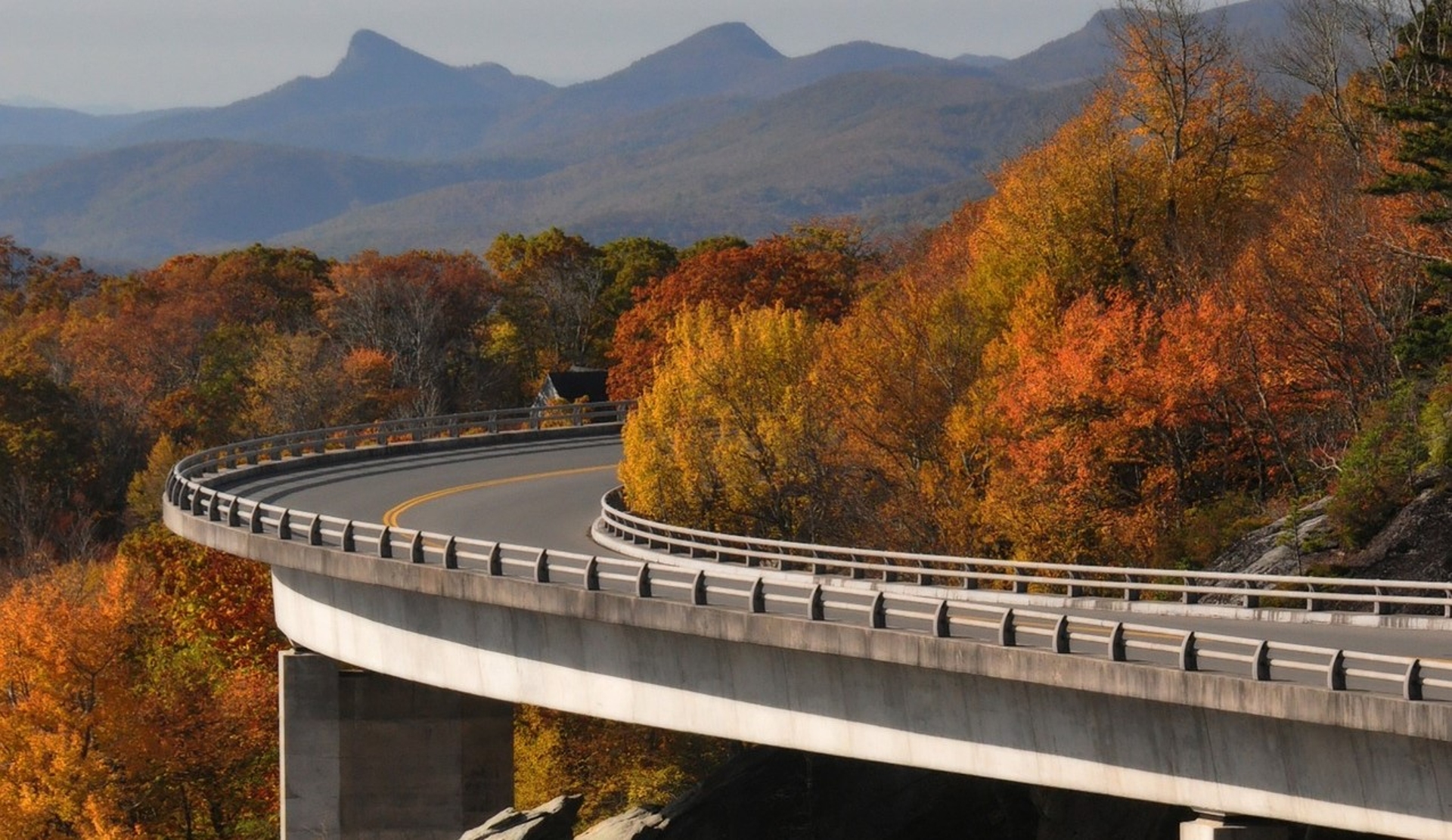 A scenic autumn view of a winding elevated roadway, flanked by colorful fall foliage, with mountain backdrops under a partly cloudy sky, beckons road trip adventures.