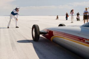 A pilot gearing up next to a streamlined land speed record car on a salt flat, while spectators and photographers observe from a distance.
