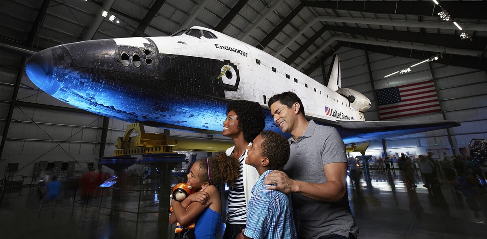 A family marvels at the impressive sight of a space shuttle exhibit in Los Angeles, with awe and curiosity reflecting on their faces as they stand beneath the spacecraft's imposing presence.