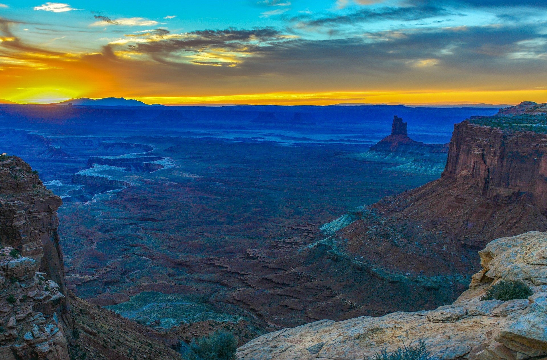 Sunset over a rugged canyon with layers of warm and cool hues painting the expansive view under a dramatic sky.