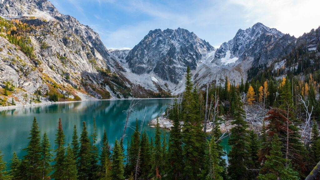 Majestic mountain peaks tower above a serene alpine lake, with evergreen and autumn-hued trees framing the tranquil waters.