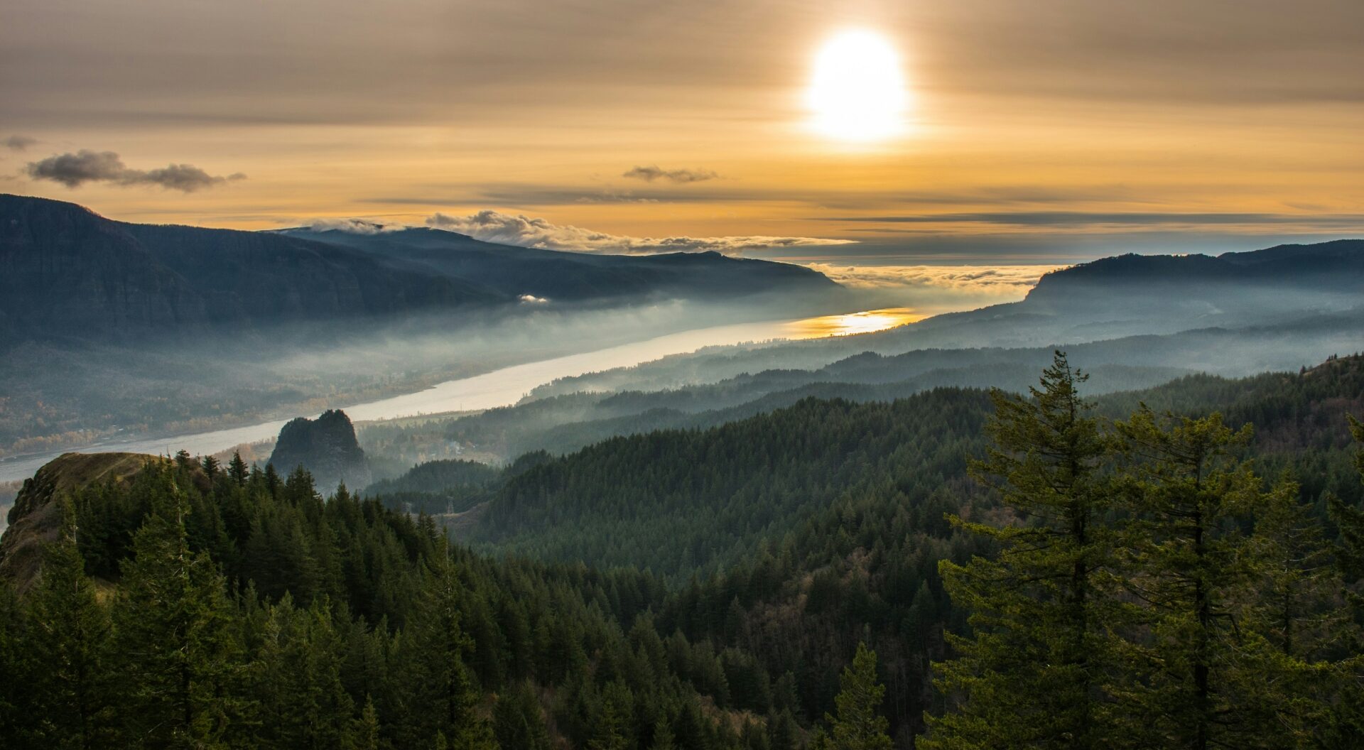 A serene sunrise illuminates a peaceful valley with misty clouds nestled between lush, rolling hills and mountains.