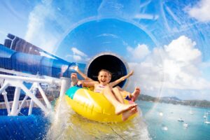 A joyful girl on a water slide, splashing down with enthusiasm, against a backdrop of blue skies and coastal scenery.