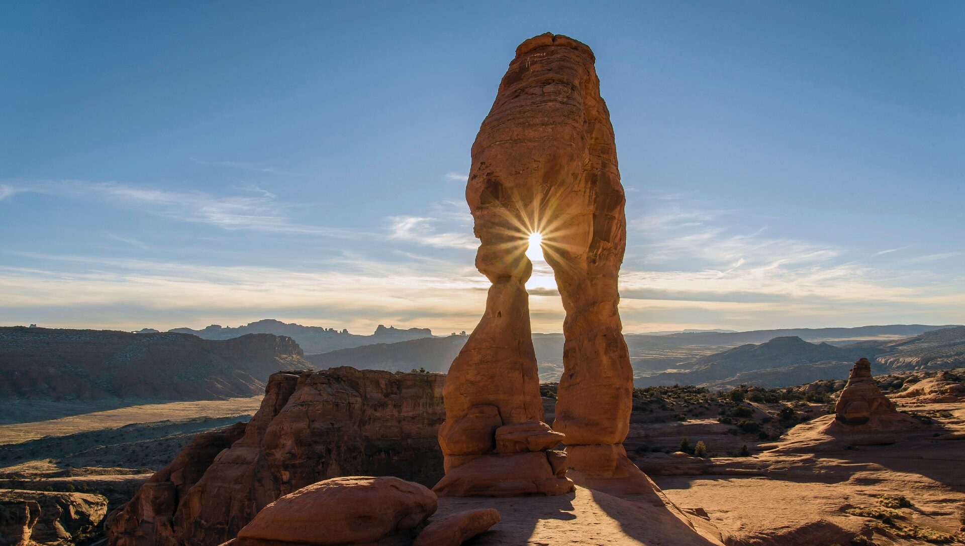 Golden rays of sunlight peek through the delicate arch, casting a warm glow on the red rock formations against the backdrop of a clear blue sky in arches national park.