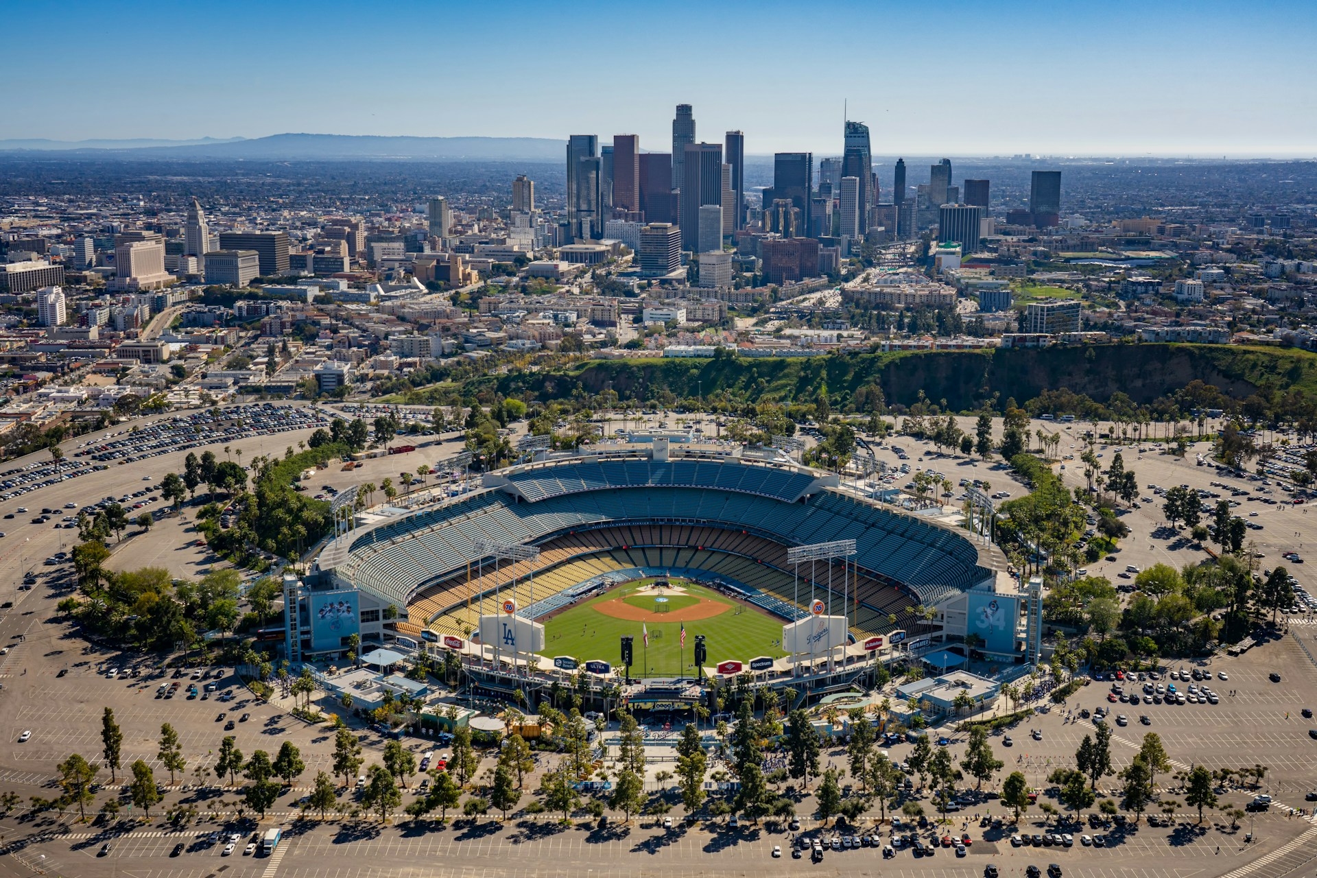 Aerial view of a Los Angeles baseball stadium in the foreground with a sprawling downtown skyline in the distance on a clear day.