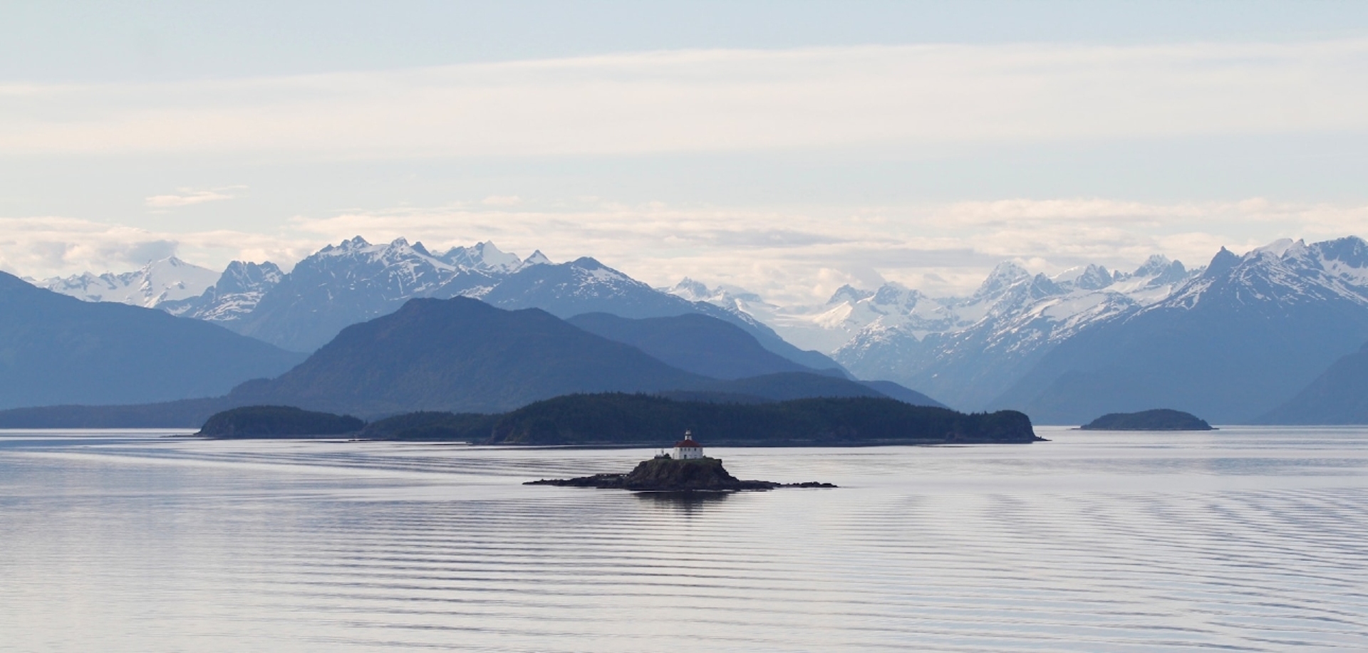 A solitary lighthouse perched on a small island with a backdrop of majestic snow-capped mountains and calm, reflective waters.