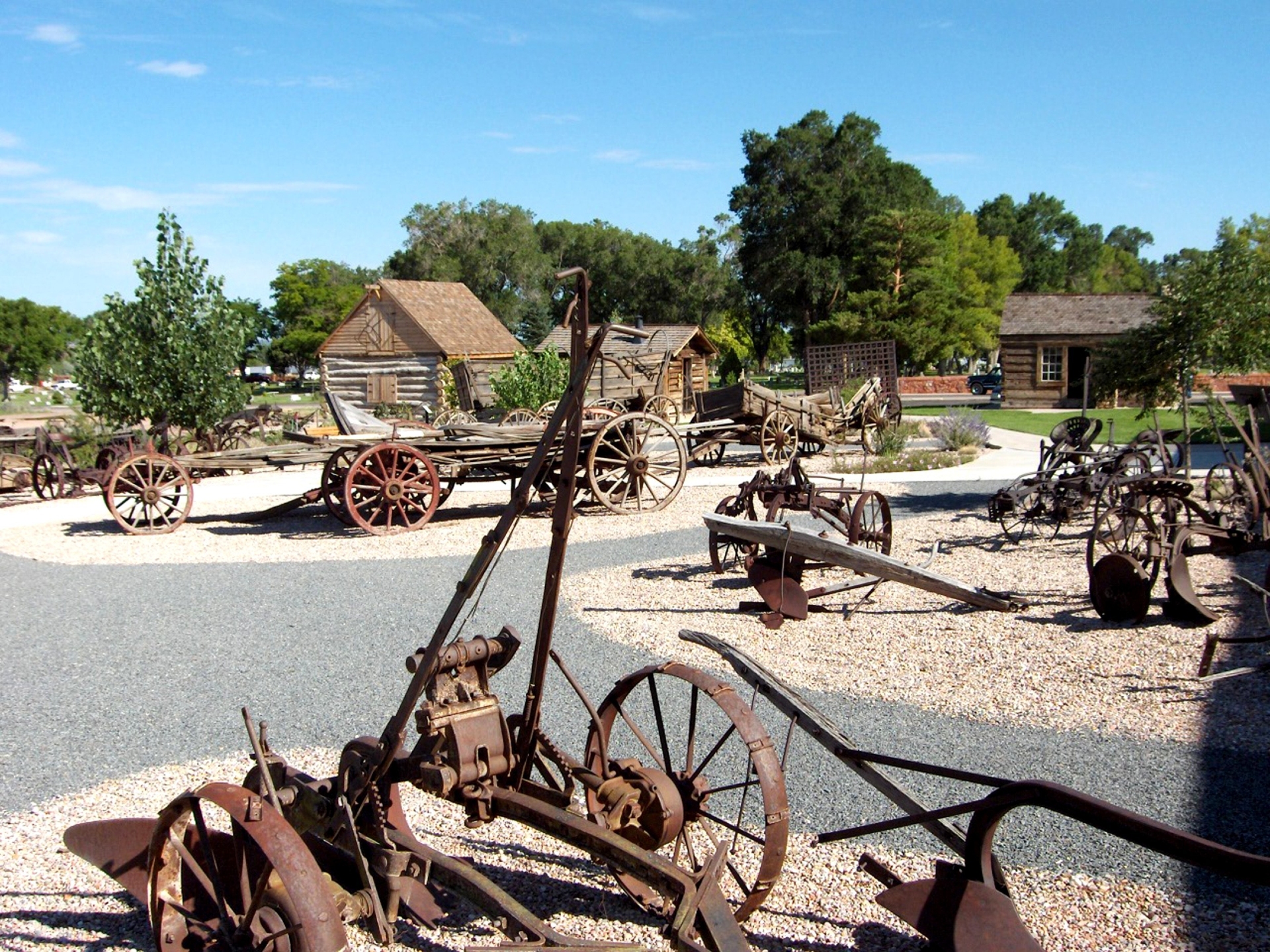 An outdoor exhibit of rustic antique farming equipment displayed in front of historical wooden cabins, evoking a bygone era.