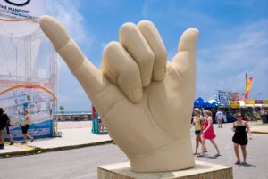 A giant sculpture of a hand making the 'shaka' sign, with people milling about in a sunny outdoor setting with carnival attractions in the background.