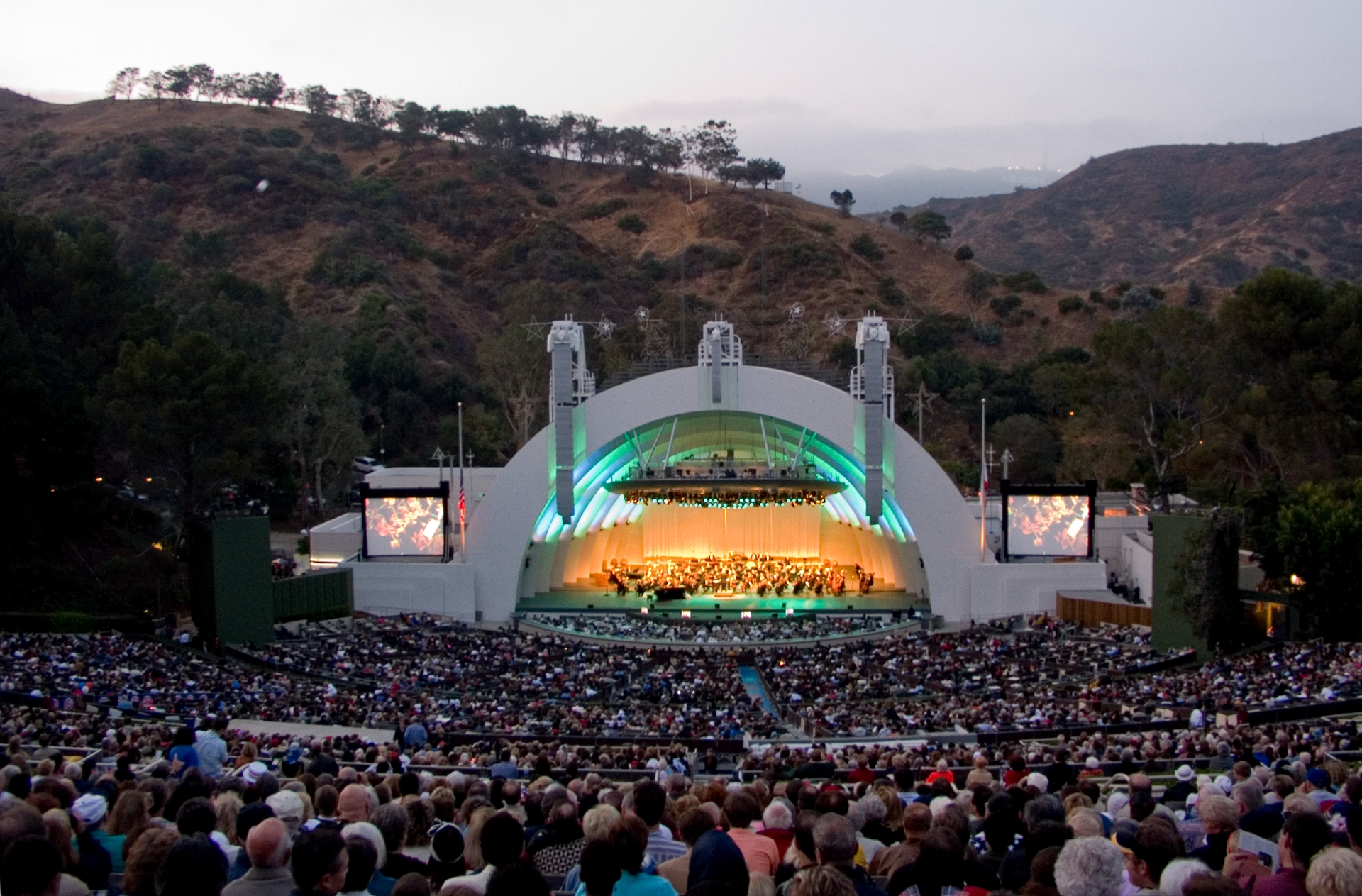An outdoor concert in full swing at a large amphitheater in Los Angeles, with a densely packed audience enjoying a live performance under a twilight sky.