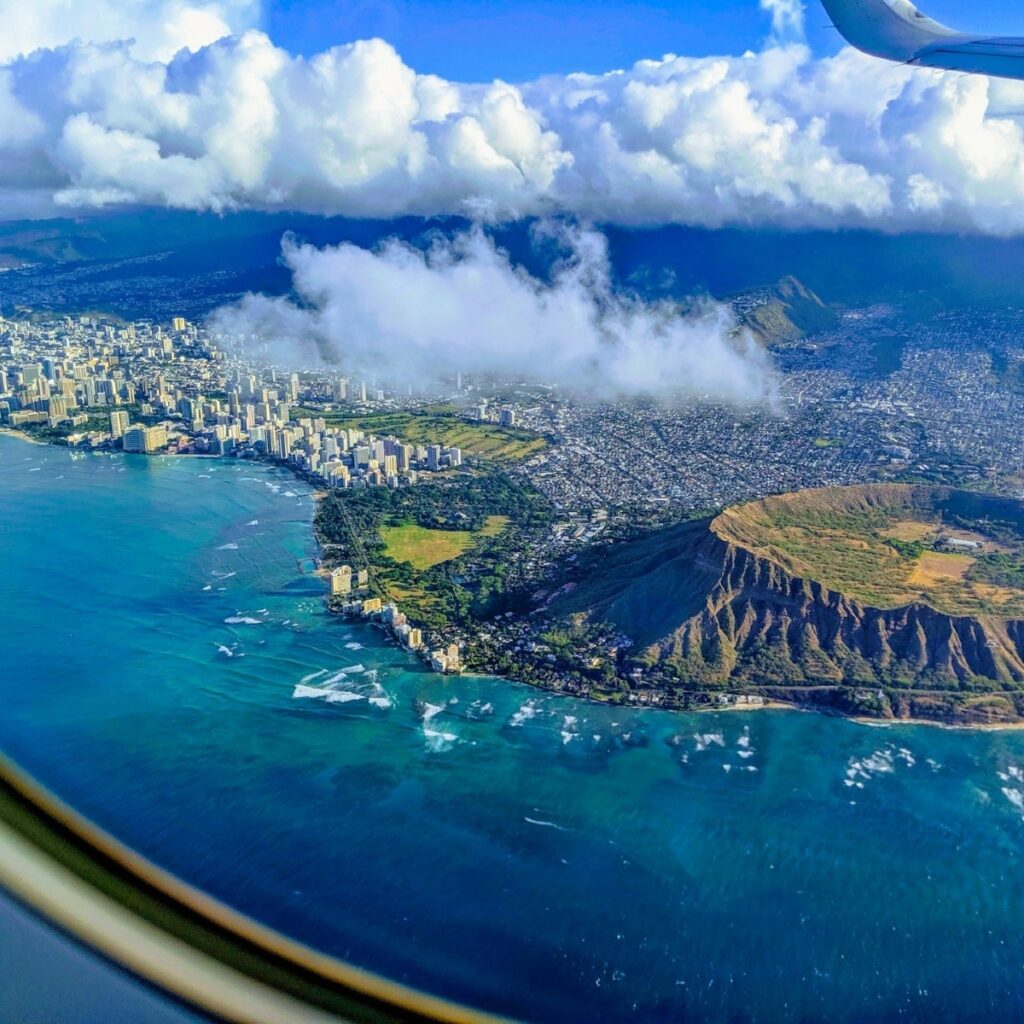An aerial view from an airplane window revealing a stunning coastal cityscape, with skyscrapers nestled against a backdrop of mountains and the vast ocean, all under the watchful eye of a beautifully sculpted cloud skyline.