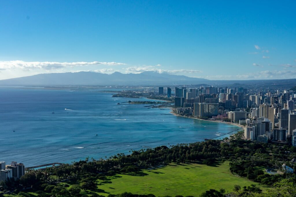 Panoramic view of a coastal city skyline with high-rise buildings along the beach, a sprawling green park in the foreground, and a mountain range in the distance under a clear blue sky.