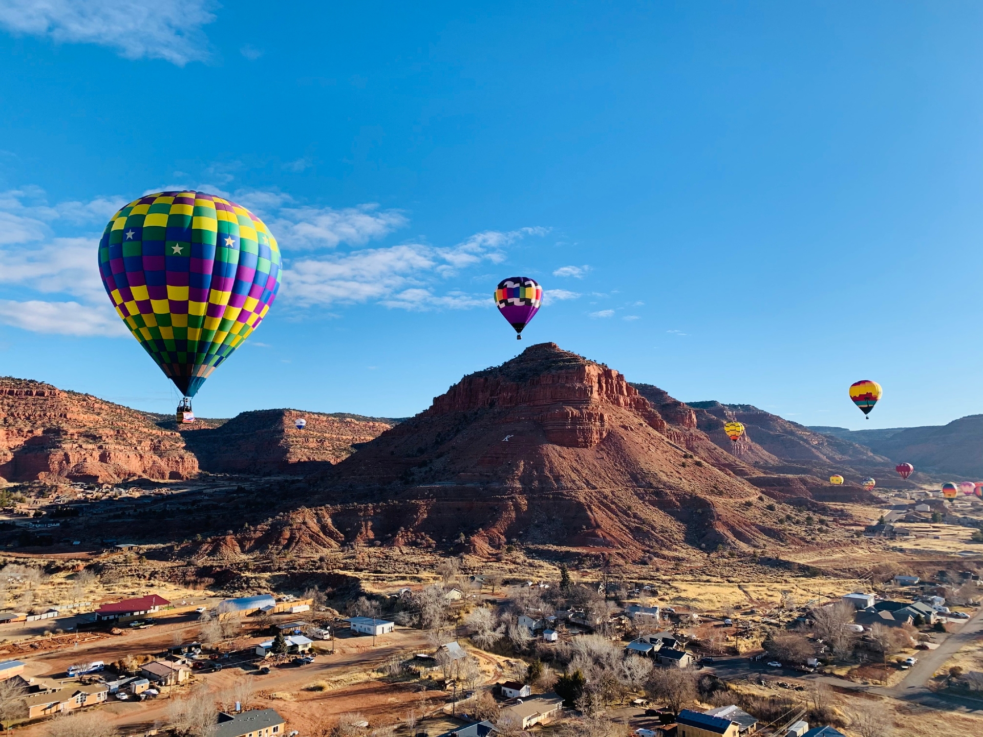 A vibrant spectacle of hot air balloons rising above a rustic village with scenic red rock formations in the background under a clear blue sky.
