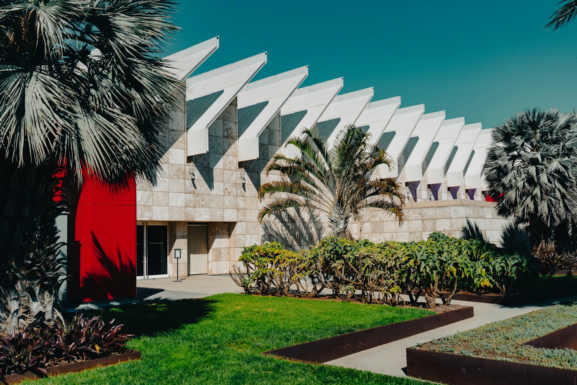 Modern architectural building in Los Angeles with distinctive white geometric patterns under a clear blue sky, complemented by lush green landscaping and a striking red entrance.