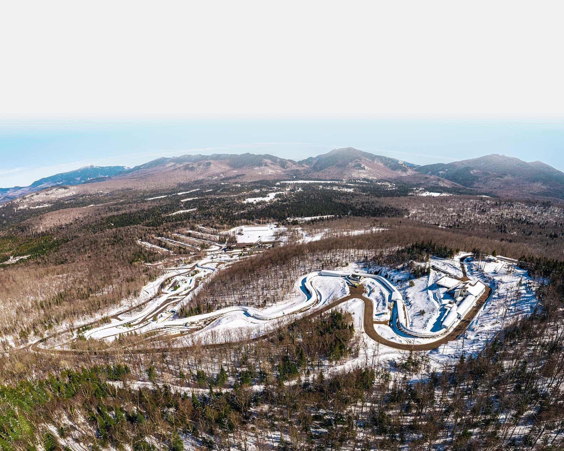 A panoramic view of a mountainous winter sports complex with winding alpine ski slopes and bobsled tracks amidst a partially snow-covered landscape, catering to adventure travel enthusiasts and thrill seekers.