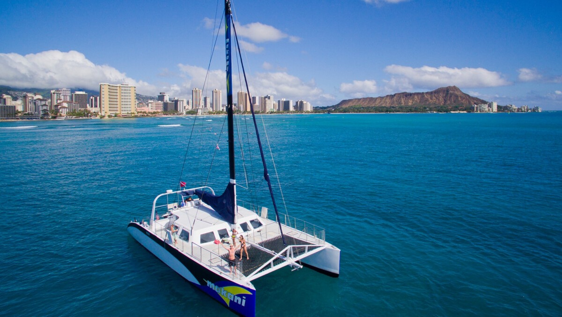 A catamaran glides over the turquoise waters off the coast of a vibrant cityscape, with the iconic ridge of a mountain in the distance under a clear blue sky.