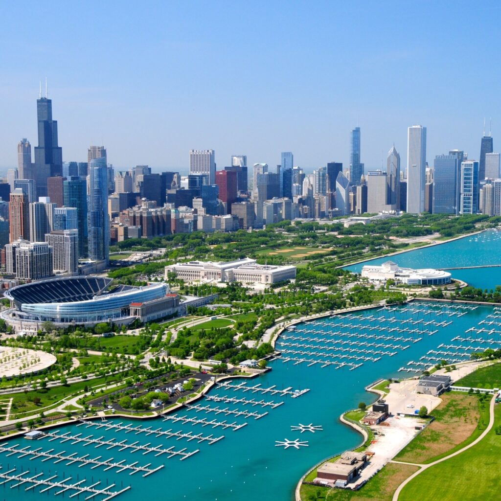 Aerial view of a vibrant city skyline with towering skyscrapers, a sports stadium, lush green parks, and a marina filled with boats beside a tranquil blue body of water.