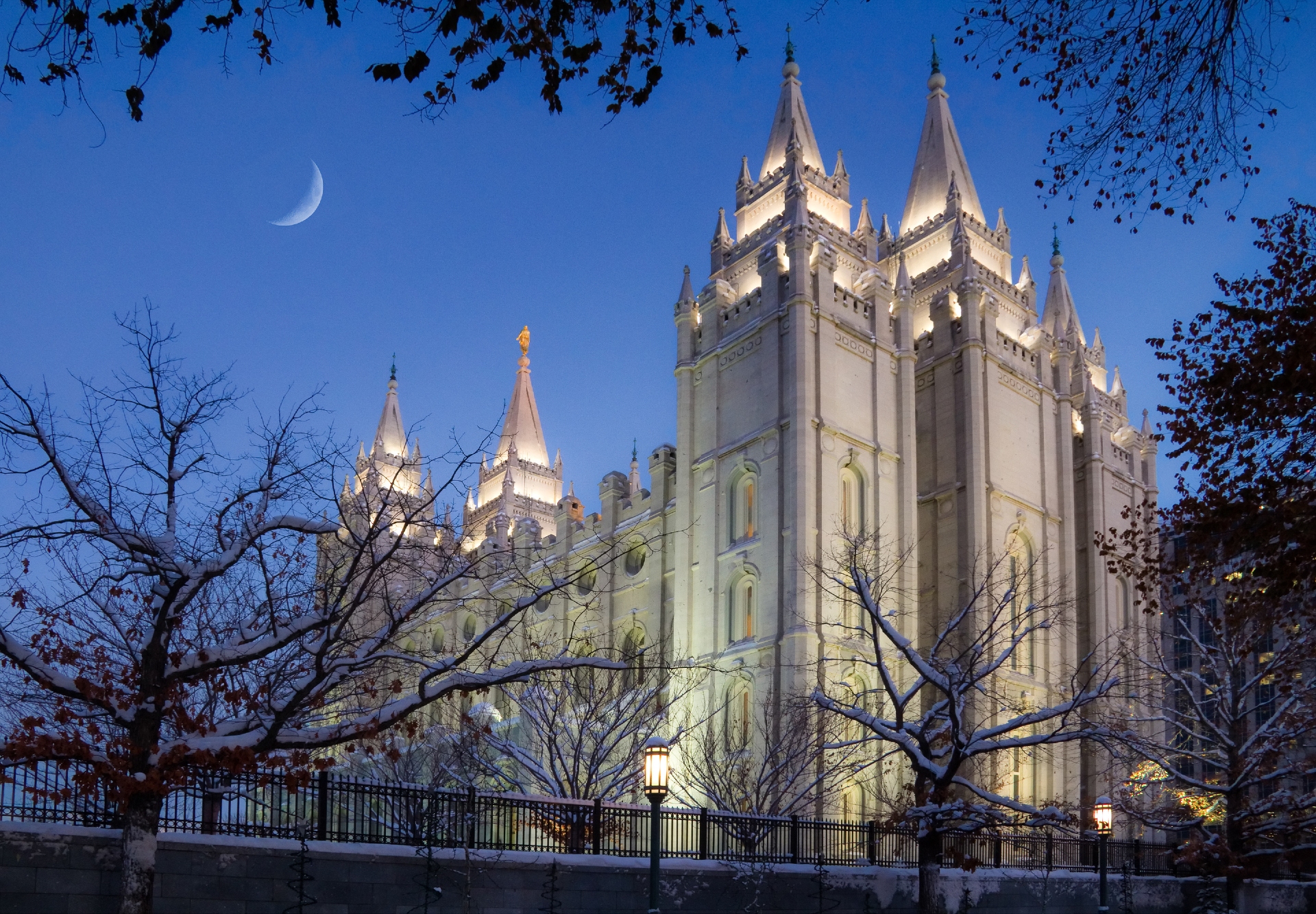 A majestic twilight view of a grand, gothic-style building with spires, illuminated against the evening sky, with a crescent moon hanging above.