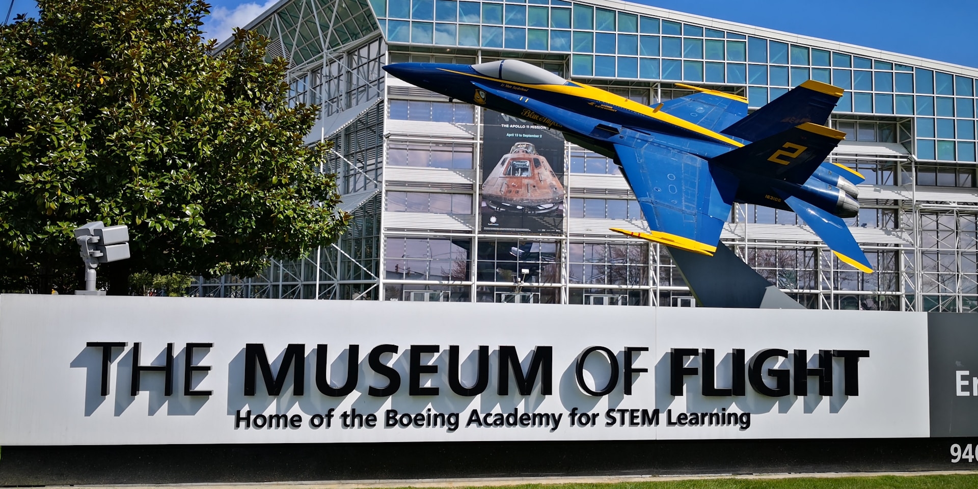 A dynamic display at the museum of flight featuring a jet fighter on a pedestal with the museum entrance and a space exhibit in the background, under a clear blue sky.