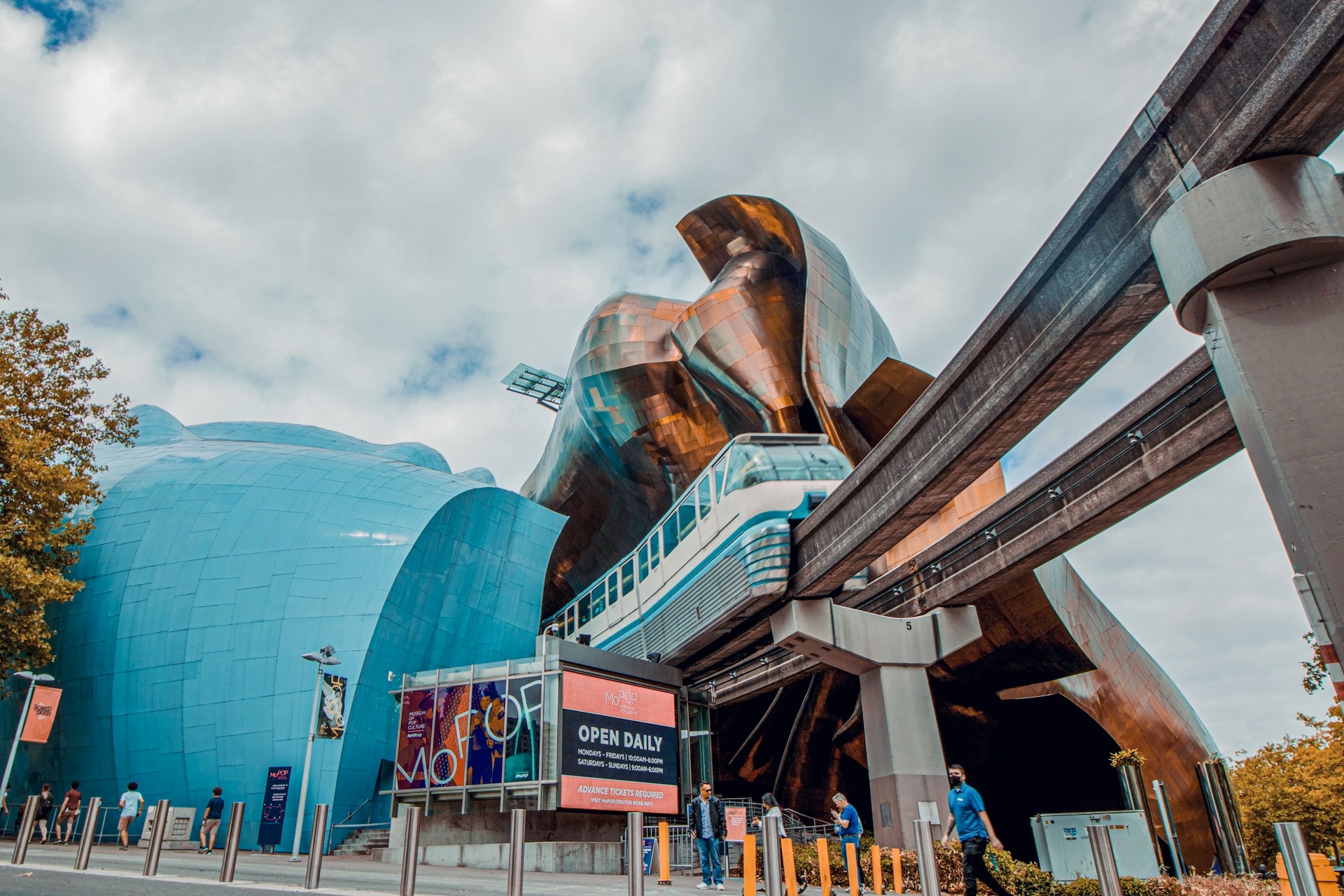 A futuristic-looking train glides through the heart of a vibrant cityscape, passing by an iconic, modern building with reflective and undulating metallic panels under a cloudy sky.