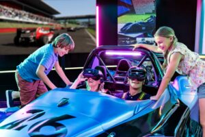 A family enjoys an immersive virtual racing experience, with two kids eagerly watching as their parents wear vr headsets in a simulated race car cockpit.