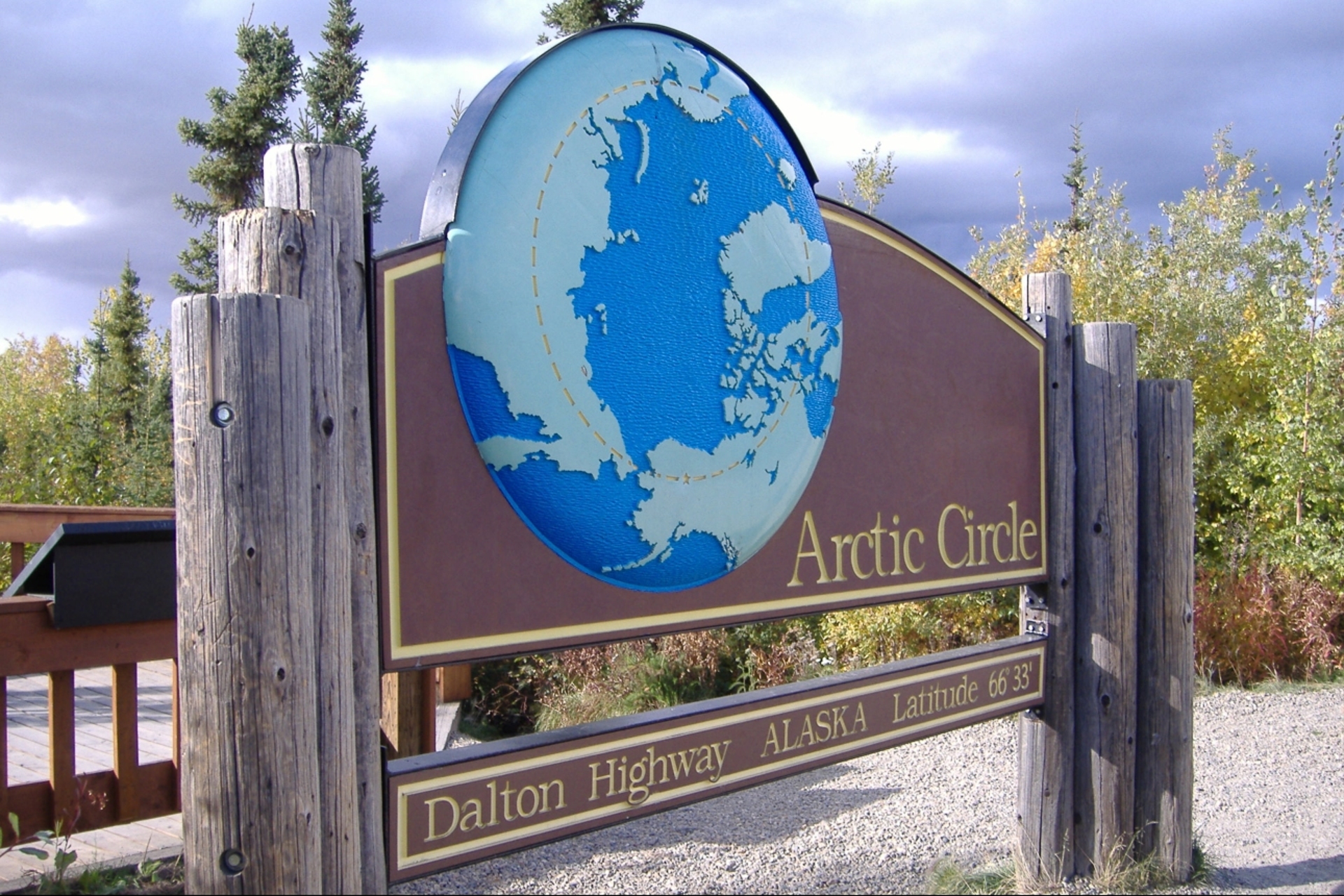 A wooden sign marking the arctic circle at latitude 66° 33′ along the dalton highway in alaska, surrounded by verdant bushes and trees under a partly cloudy sky.