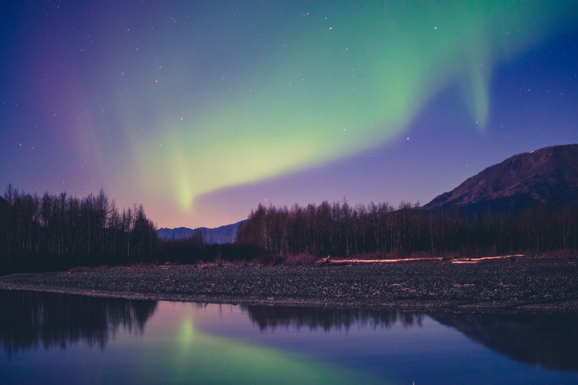 A tranquil night scene with the vibrant aurora borealis dancing across the starlit sky, reflecting on the serene surface of a still river, bordered by a silhouette of a forest and mountains.