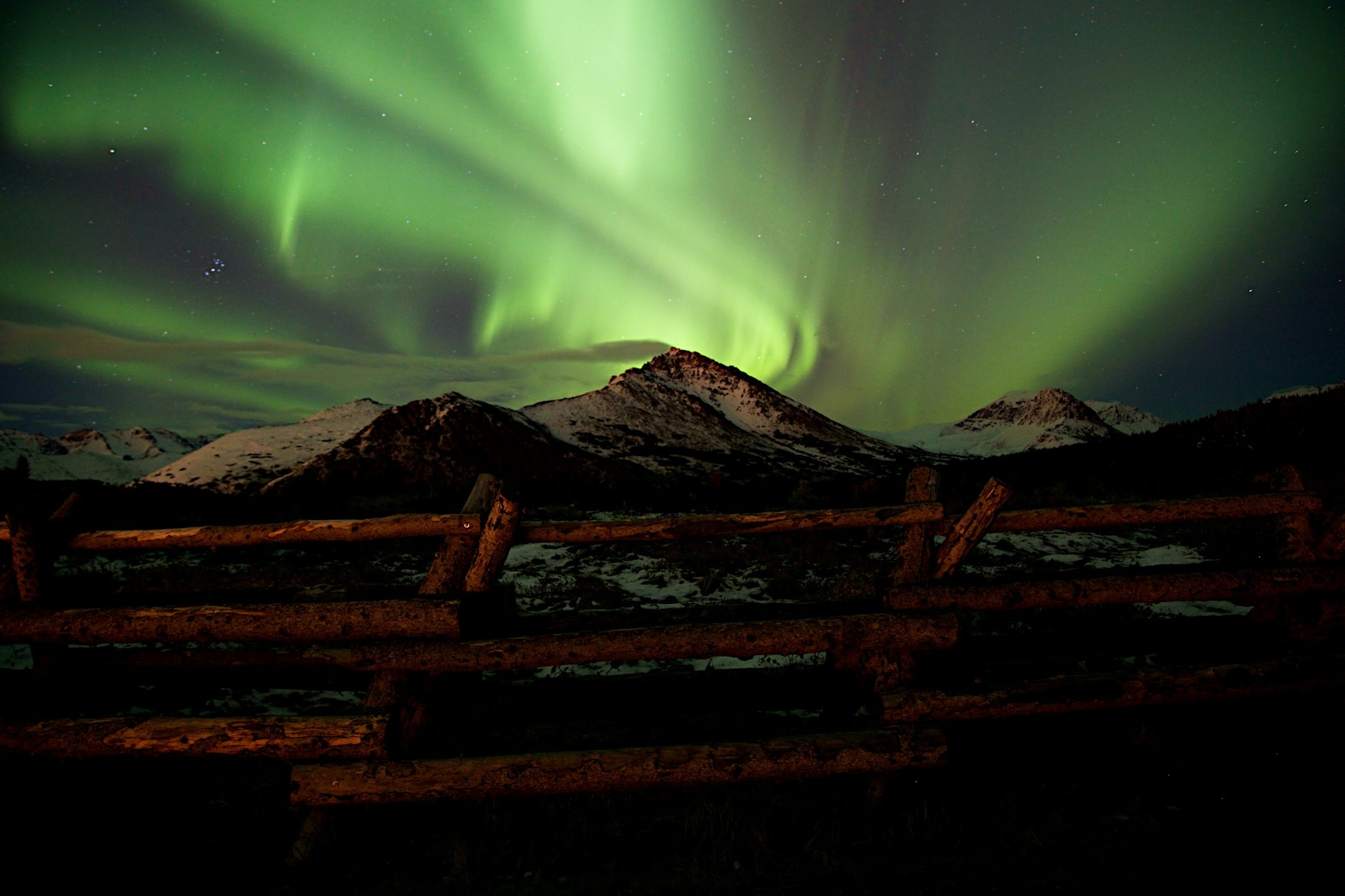 A stunning display of the northern lights (aurora borealis) illuminating the night sky with vibrant green hues above snow-capped mountains, with a rustic wooden fence in the foreground.