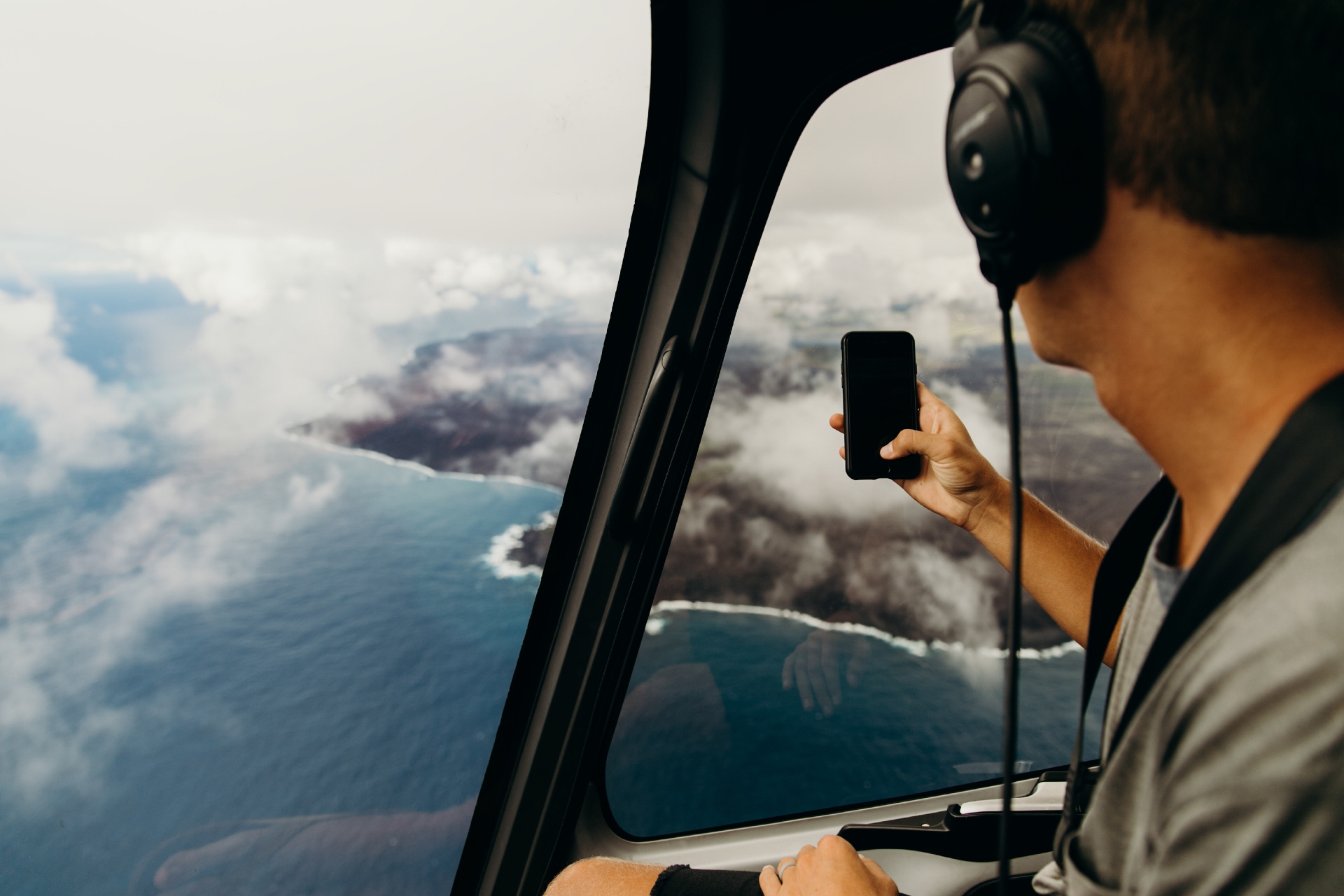 An individual wearing headphones captures a stunning aerial view of a coastline and clouds through the window of an aircraft using a smartphone.
