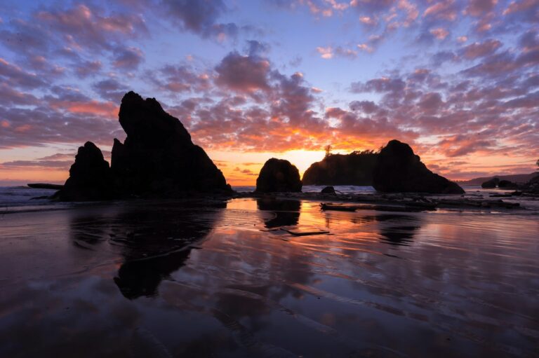 A majestic sunset silhouettes towering sea stacks against a vibrant sky, reflected on the serene, wet sand of a tranquil beach.