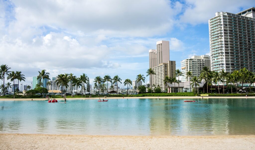 A serene tropical lagoon with people enjoying paddleboarding, bordered by a sandy beach and flanked by tall palm trees and modern buildings under a partly cloudy sky.