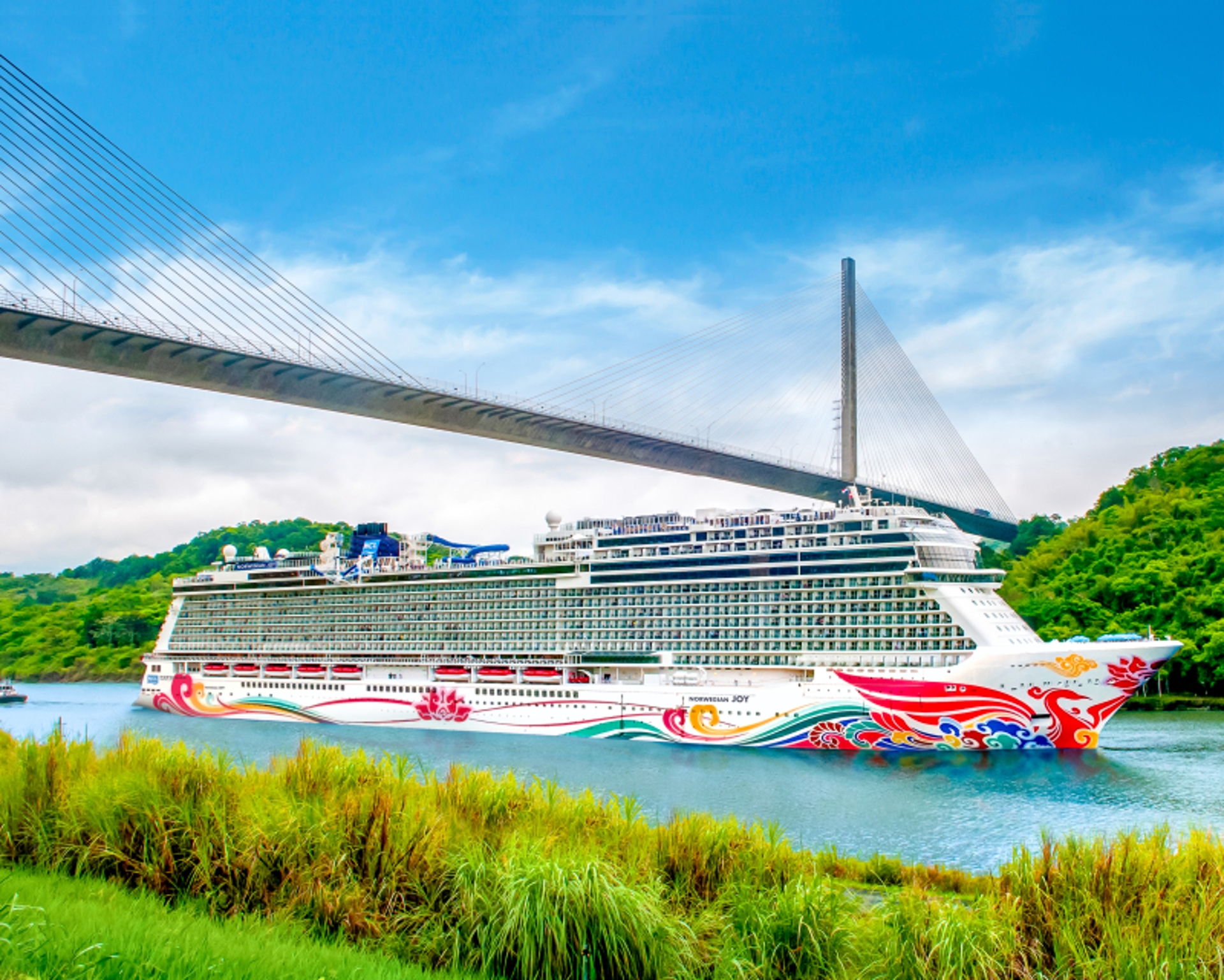 A vibrant cruise ship adorned with colorful artwork embarks on memorable voyages beneath a modern suspension bridge, set against a backdrop of lush green hills and a bright blue sky with scattered clouds.