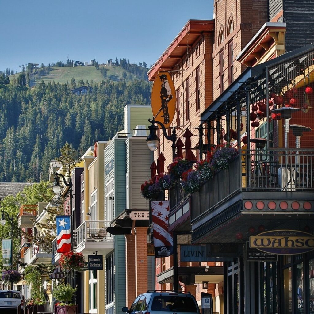 Charming mountain town street lined with historic buildings, featuring balconies adorned with colorful flowers and a backdrop of lush green hills.