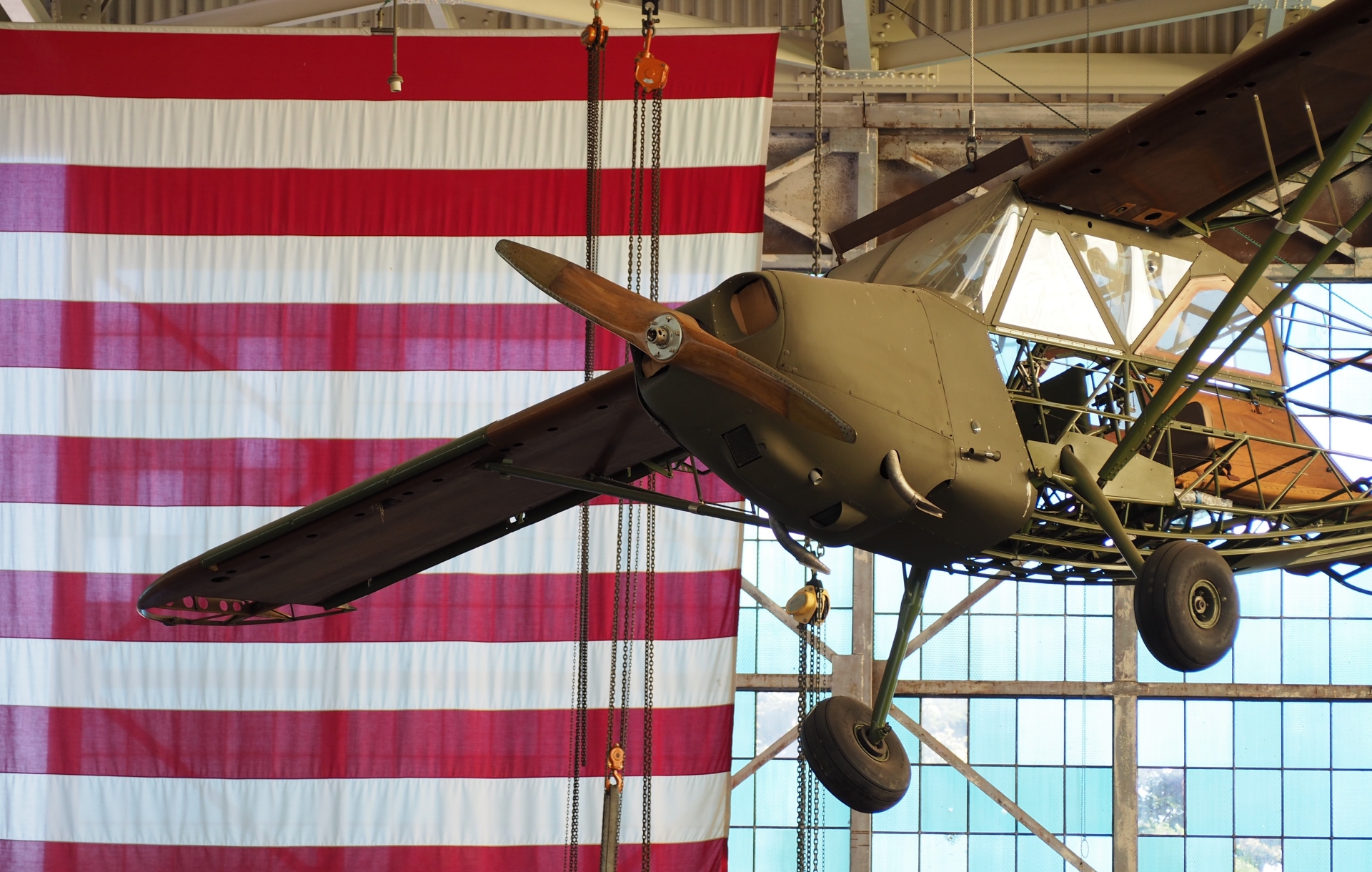 A vintage biplane suspended in front of a large american flag, displayed as a piece of aviation history in an exhibition setting.