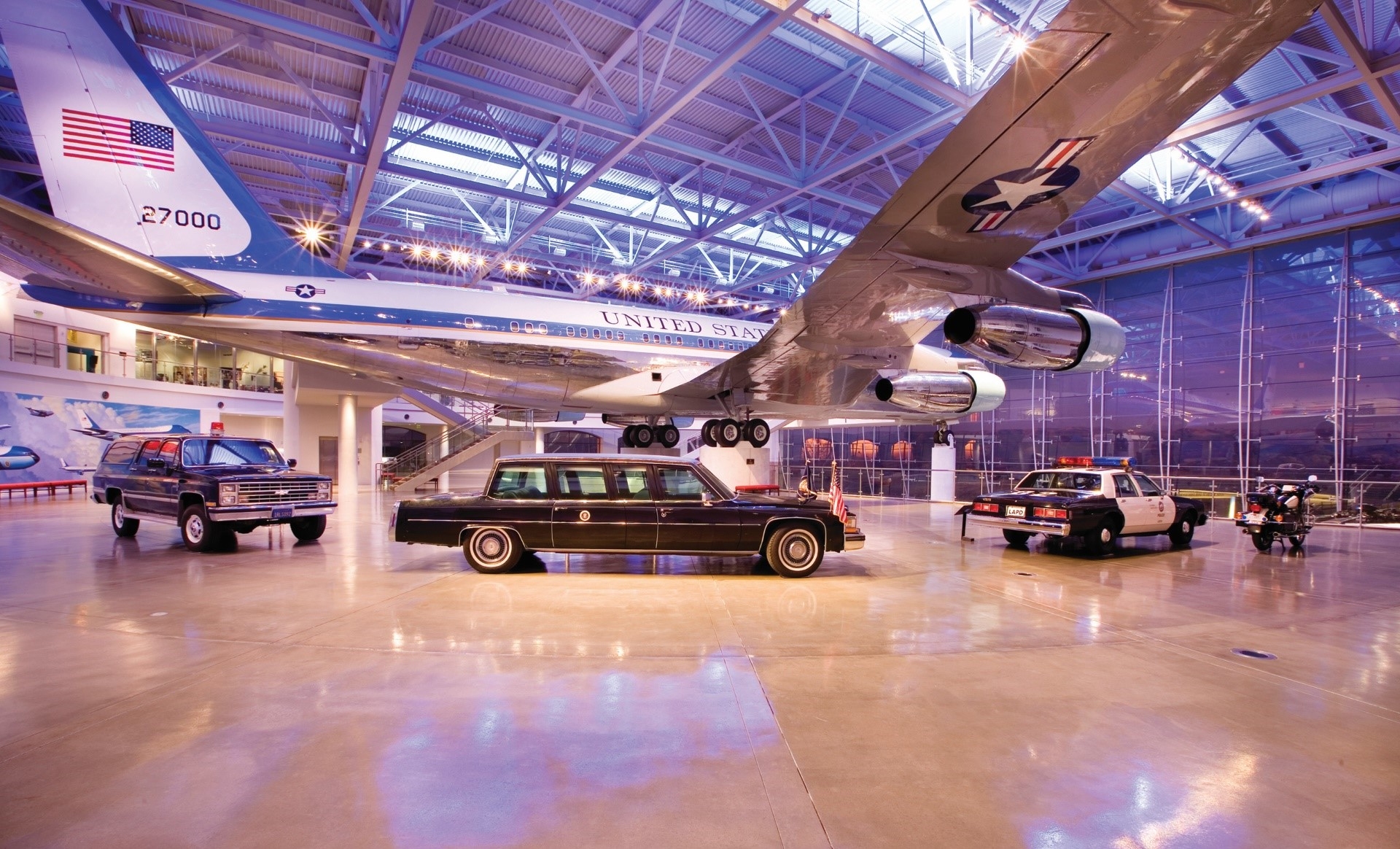 A spacious hangar in Los Angeles showcasing an iconic air force one aircraft flanked by historical vehicles, all exhibited under a transparent roof.